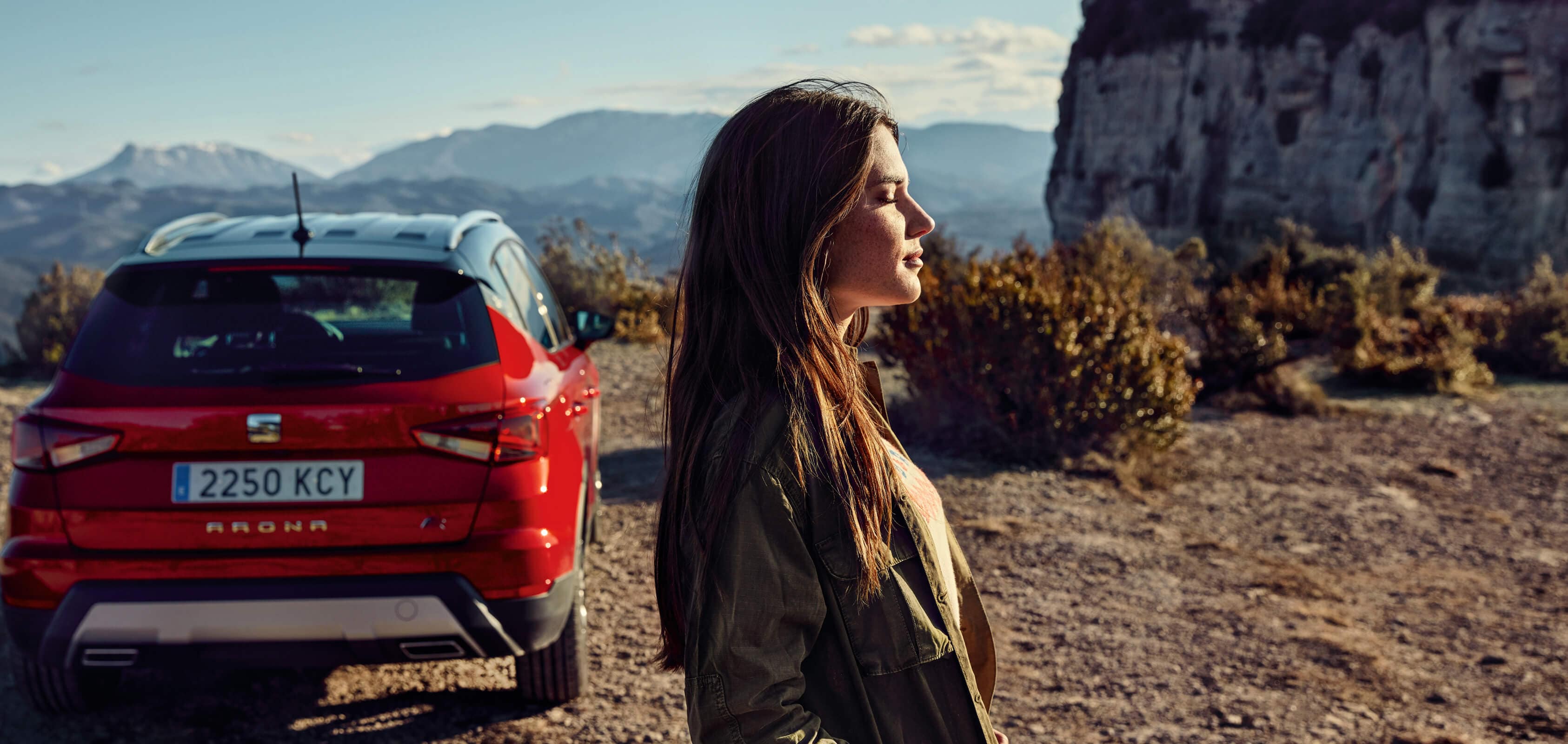 SEAT new car services – rear view of SEAT Arona crossover SUV parked in a dessert with a profile view of a woman